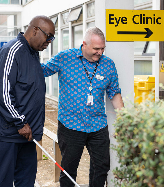 An eye clinic liaison officer (ECLO) guiding a man with a cane into a hospital eye clinic.